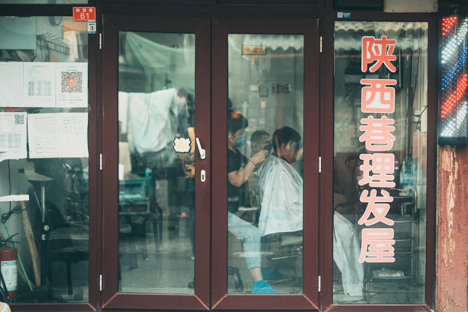 The image shows a hair salon with large windows that reflect the interior, creating an interesting visual effect. The salon is filled with various hair products and tools, indicating it's a professional setting for hairdressing services. Two people are seated in chairs inside the salon, one of whom appears to be cutting another person's hair using scissors. A sign on the window displays Chinese characters that translate to "hair salon".