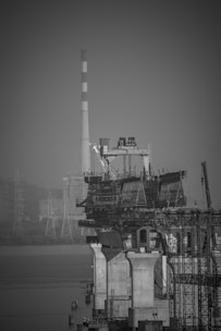 A black and white photograph of a bridge under construction over a body of water. The scene includes large concrete pillars supporting partially built sections of the bridge. In the background, there is an industrial chimney with stripes, and several towers, possibly for electrical lines, are visible against a hazy sky.