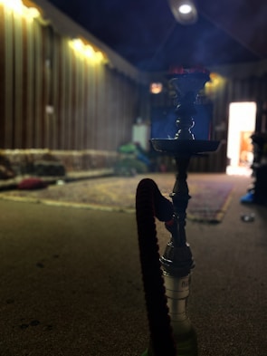 A traditional setting with dim lighting features a hookah placed prominently on the carpeted floor. In the background, the room is furnished with cushions and blankets, giving a cozy and relaxed ambiance. Light filters through a partially open door.