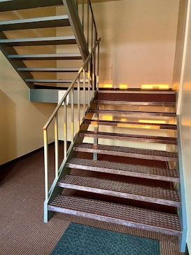 A metal staircase inside a building, with natural light streaming in, creating patterns on the steps and walls. The stairs have a textured surface and a sturdy railing. The floor is carpeted with a brown pattern, and a small green mat is placed at the base of the stairs.