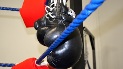 A pair of black boxing gloves is hanging on the blue ropes of a boxing ring. The ropes are lined with red protective padding. The background is a light-colored wall, possibly within a gym.