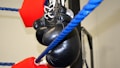 A pair of black boxing gloves is hanging on the blue ropes of a boxing ring. The ropes are lined with red protective padding. The background is a light-colored wall, possibly within a gym.