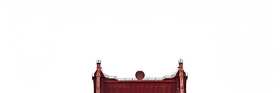 The upper section of a red iron structure, resembling a bridge or industrial edifice, is visible against a plain white background. The structure features vertical and horizontal beams with small finial-like ornaments on top.