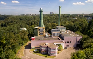 An industrial facility with a large chimney surrounded by dense forest. The structure has multiple buildings with flat rooftops and interconnected pipes. The sky is clear with a few clouds, and in the distance, some city buildings are visible.
