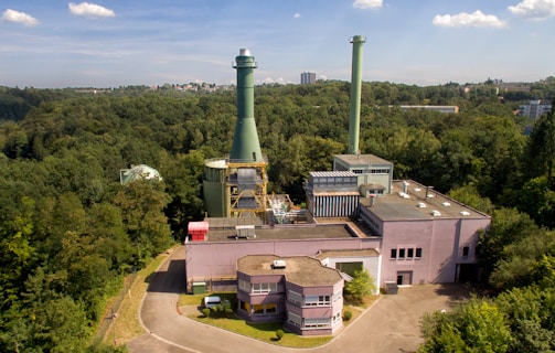 An industrial facility with a large chimney surrounded by dense forest. The structure has multiple buildings with flat rooftops and interconnected pipes. The sky is clear with a few clouds, and in the distance, some city buildings are visible.