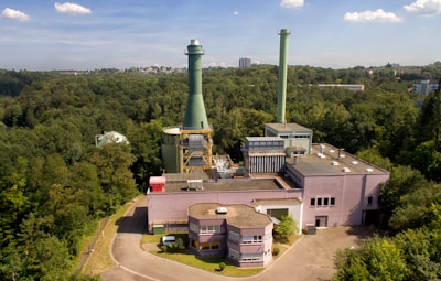 An industrial facility with a large chimney surrounded by dense forest. The structure has multiple buildings with flat rooftops and interconnected pipes. The sky is clear with a few clouds, and in the distance, some city buildings are visible.