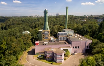 An industrial facility with a large chimney surrounded by dense forest. The structure has multiple buildings with flat rooftops and interconnected pipes. The sky is clear with a few clouds, and in the distance, some city buildings are visible.