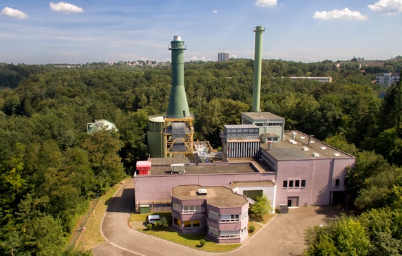 An industrial facility with a large chimney surrounded by dense forest. The structure has multiple buildings with flat rooftops and interconnected pipes. The sky is clear with a few clouds, and in the distance, some city buildings are visible.