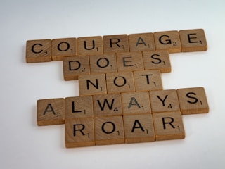 Wooden letter tiles are arranged on a flat surface to form the phrase 'COURAGE DOES NOT ALWAYS ROAR'. The tiles are reminiscent of Scrabble pieces, with each letter having a numerical value at the bottom right corner. The background is a plain, light color.