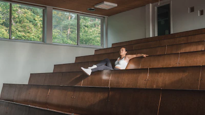 A person is seated alone in a large, empty lecture hall with wooden benches. They are dressed in casual athletic wear, including a white top and dark leggings, and appear to be relaxed with one arm resting on the back of the bench. Large windows in the background reveal lush greenery outside, indicating a connection to nature.