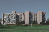 A large, multi-story residential building with numerous balconies is set against a clear blue sky. A grassy area and path are in the foreground, with two people interacting — one walking and the other on a bicycle.