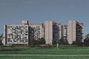 A large, multi-story residential building with numerous balconies is set against a clear blue sky. A grassy area and path are in the foreground, with two people interacting — one walking and the other on a bicycle.