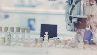 A laboratory setting with a robotic arm interacting with test tubes that have various colored caps, arranged on a tray. The background has a blurred effect, creating a focus on the equipment.