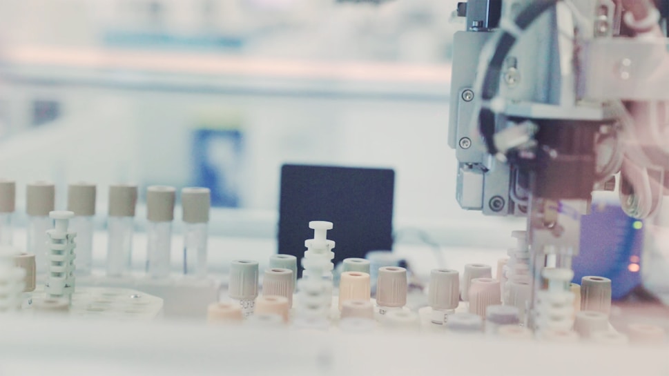 A laboratory setting with a robotic arm interacting with test tubes that have various colored caps, arranged on a tray. The background has a blurred effect, creating a focus on the equipment.