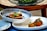 A close-up of two dishes artfully arranged. The first dish features a gourmet tart topped with colorful vegetables and garnished with greens. The second dish presents fried croquettes accompanied by a garnish and sauce, all arranged on elegant white plates.