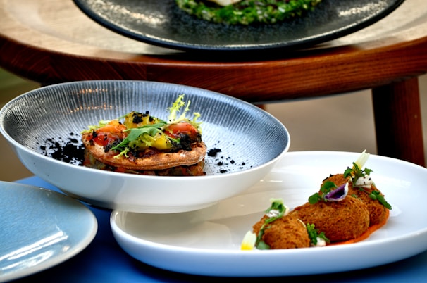 A close-up of two dishes artfully arranged. The first dish features a gourmet tart topped with colorful vegetables and garnished with greens. The second dish presents fried croquettes accompanied by a garnish and sauce, all arranged on elegant white plates.
