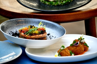 A close-up of two dishes artfully arranged. The first dish features a gourmet tart topped with colorful vegetables and garnished with greens. The second dish presents fried croquettes accompanied by a garnish and sauce, all arranged on elegant white plates.
