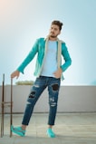 A young man with styled hair is casually standing on a tiled surface. He's wearing a teal jacket with a white fur collar over a white t-shirt, distressed jeans with large rips, and matching teal shoes with patterns. One of his hands is resting on a metal rod structure, and the background shows an outdoor setting under a clear sky.