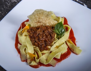 A plate of wide ribbon pasta topped with a rich, red tomato or meat-based sauce. The dish is garnished with a fresh green herb, possibly basil, and a crispy, golden-brown textured piece, likely cheese or breadcrumb. Bright yellow flower petals add a touch of color and elegance.