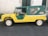 A vintage vehicle with a contrasting yellow and green color scheme is parked on a dark asphalt surface. It features a convertible-like design with a soft green top and clear plastic windows. The vehicle's body is yellow with a ribbed texture, and the wheels are white.