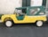 A vintage vehicle with a contrasting yellow and green color scheme is parked on a dark asphalt surface. It features a convertible-like design with a soft green top and clear plastic windows. The vehicle's body is yellow with a ribbed texture, and the wheels are white.