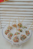 Colorful cake pops coated with vibrant rainbow sprinkles are arranged neatly on a glass stand. Each cake pop is placed in a white paper cup and skewered with a thin stick. The background consists of a light, wooden panel texture, adding a simple and clean aesthetic to the setup.