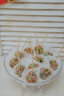 Colorful cake pops coated with vibrant rainbow sprinkles are arranged neatly on a glass stand. Each cake pop is placed in a white paper cup and skewered with a thin stick. The background consists of a light, wooden panel texture, adding a simple and clean aesthetic to the setup.