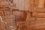 A close-up view of a large rusted metal structure with intricate, layered details. The structure incorporates industrial elements such as rivets, machine parts, and a chair. The surface is heavily corroded, giving it a rough, textured appearance. Lines and geometric shapes are prevalent throughout the design, giving a sense of mechanical complexity.