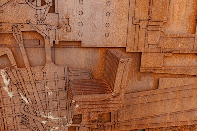 A close-up view of a large rusted metal structure with intricate, layered details. The structure incorporates industrial elements such as rivets, machine parts, and a chair. The surface is heavily corroded, giving it a rough, textured appearance. Lines and geometric shapes are prevalent throughout the design, giving a sense of mechanical complexity.