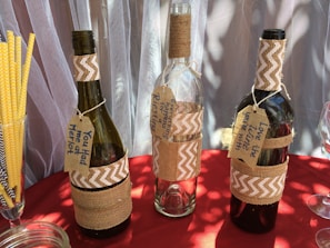 Three wine bottles wrapped in burlap and decorated with chevron-patterned fabric are displayed. Each bottle features a handmade tag with playful messages related to wine. Yellow striped straws and a glass jar are seen alongside on a red tablecloth. The backdrop consists of sheer curtains.
