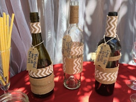 Three wine bottles wrapped in burlap and decorated with chevron-patterned fabric are displayed. Each bottle features a handmade tag with playful messages related to wine. Yellow striped straws and a glass jar are seen alongside on a red tablecloth. The backdrop consists of sheer curtains.