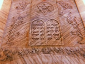 A carved stone plaque features two tablets with Hebrew inscriptions. At the top is a Star of David with the word 'אמת' (truth) inside it. The surrounding design includes floral and ornamental motifs intricately carved into the stone.