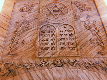 A carved stone plaque features two tablets with Hebrew inscriptions. At the top is a Star of David with the word 'אמת' (truth) inside it. The surrounding design includes floral and ornamental motifs intricately carved into the stone.