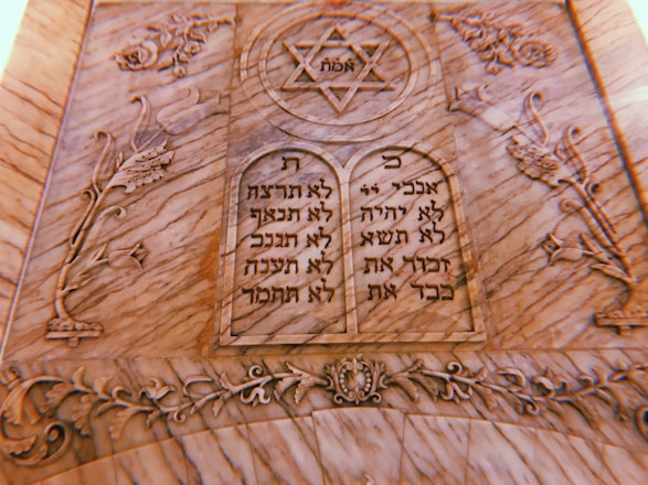 A carved stone plaque features two tablets with Hebrew inscriptions. At the top is a Star of David with the word 'אמת' (truth) inside it. The surrounding design includes floral and ornamental motifs intricately carved into the stone.
