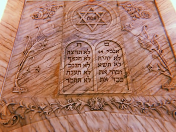 A carved stone plaque features two tablets with Hebrew inscriptions. At the top is a Star of David with the word 'אמת' (truth) inside it. The surrounding design includes floral and ornamental motifs intricately carved into the stone.