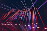 Bright colored laser beams in red and blue cut across a darkened room, originating from a stage with a DJ booth that displays a logo. The beams reflect off the glossy floor, creating a dynamic and energetic atmosphere.
