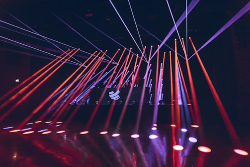 Bright colored laser beams in red and blue cut across a darkened room, originating from a stage with a DJ booth that displays a logo. The beams reflect off the glossy floor, creating a dynamic and energetic atmosphere.