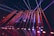 Bright colored laser beams in red and blue cut across a darkened room, originating from a stage with a DJ booth that displays a logo. The beams reflect off the glossy floor, creating a dynamic and energetic atmosphere.