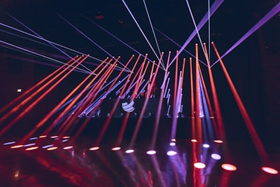 Bright colored laser beams in red and blue cut across a darkened room, originating from a stage with a DJ booth that displays a logo. The beams reflect off the glossy floor, creating a dynamic and energetic atmosphere.