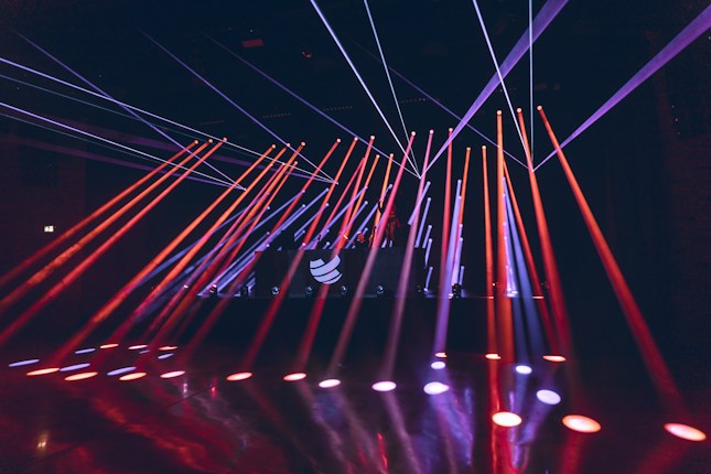 Bright colored laser beams in red and blue cut across a darkened room, originating from a stage with a DJ booth that displays a logo. The beams reflect off the glossy floor, creating a dynamic and energetic atmosphere.