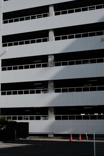 A multi-story parking garage with a modern design featuring horizontal lines and dark window slits. The white facade creates a minimalistic look, with visible shadows adding depth. A couple of traffic cones are positioned near the entrance at the bottom.