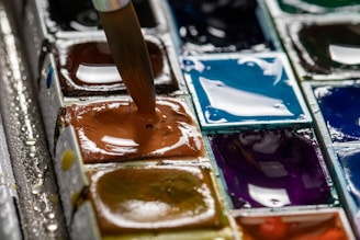 A close-up of a watercolor paint palette featuring a variety of wet paint pans. A brush is being dipped into one of the paint pans, which is a rich brown hue. The other colors visible include shades of blue, purple, and green, with a glossy and wet surface appearance.