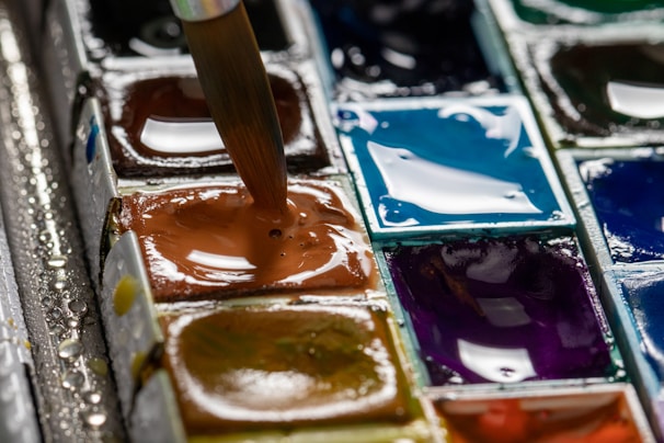 A close-up of a watercolor paint palette featuring a variety of wet paint pans. A brush is being dipped into one of the paint pans, which is a rich brown hue. The other colors visible include shades of blue, purple, and green, with a glossy and wet surface appearance.