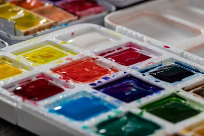 A close-up view of a palette filled with various vibrant watercolor paints. The paint is arranged in small, square compartments, showcasing a wide range of colors including reds, yellows, blues, greens, and more. The surface of the paints appears wet and glossy, suggesting they have been recently used.