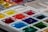 A close-up view of a palette filled with various vibrant watercolor paints. The paint is arranged in small, square compartments, showcasing a wide range of colors including reds, yellows, blues, greens, and more. The surface of the paints appears wet and glossy, suggesting they have been recently used.