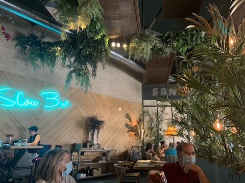 A cozy cafe interior with a wooden herringbone wall design is adorned with lush green plants hanging from the ceiling. The neon sign 'Slow Bar' is illuminated in blue on the wall. Several people are seated, some wearing face masks. The atmosphere is relaxed with dim lighting from decorative lamps. Wooden tables and chairs, along with woven baskets, add to the rustic decor.