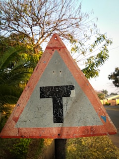 A weathered triangular road sign with a red border and a black 'T' symbol in the center, indicating a T-intersection ahead. The background shows lush greenery with bushes and trees, under a clear blue sky.