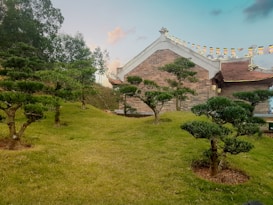 A serene garden landscape with neatly trimmed trees and lush green grass. In the background, a building with traditional architectural elements stands, adorned with colorful flags. The calm sky above adds to the tranquil atmosphere.