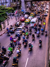 A congested urban street filled with numerous motorcycles and cars. Motorcycles dominate the foreground, each rider wearing a helmet and various colored clothing. Cars and taxis form a parallel line in the adjacent lane. Tall buildings and power lines frame the background, with a large concrete overpass stretching across the street.