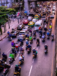 A congested urban street filled with numerous motorcycles and cars. Motorcycles dominate the foreground, each rider wearing a helmet and various colored clothing. Cars and taxis form a parallel line in the adjacent lane. Tall buildings and power lines frame the background, with a large concrete overpass stretching across the street.